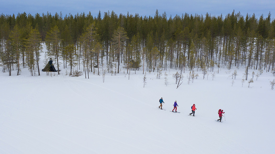 Neljä lumikenkäilijää ilmakuvassa kulkee laavulta lumisen lammen poikki.