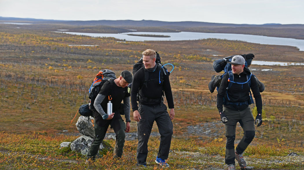 Kolme retkelijää rinkat selässä kävelemässä tunturin lakea kohti hymyssä suin. Taustalla näkyy harvaa puustoa ja vesistö.