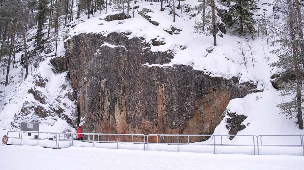 Retkeilijä katselee kallioseinämässä olevia maalauksia. Kallion äärellä on metallista tehty kävelysilta. Maassa on lunta.