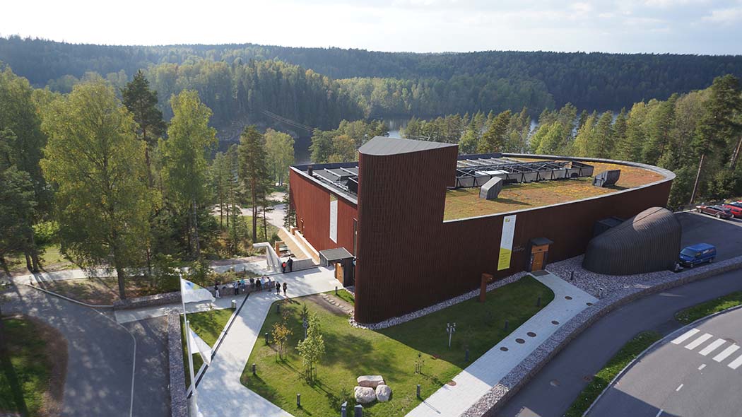 Iso pyöreäkulmainen rakennus mäen harjanteella.Edustalla auto- ja kävelyteitä. Taustalla järvi ja kumpuilevaa metsämaastoa.Rakennuksen sisäänkäynnin edustalla ihmisiä.