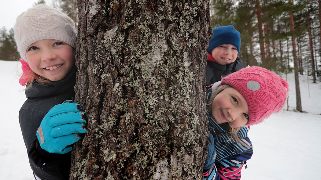 Lapset kurkkivat puun rungon takaa talvisessa metsässä.