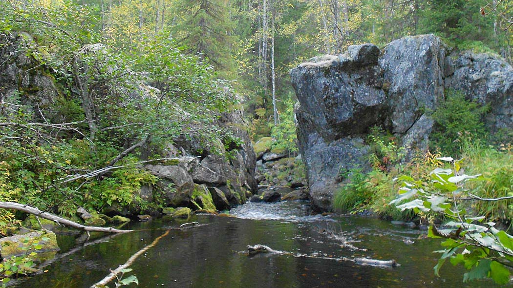 Kahden suuren kivilohkareen välissä virtaa joki. Ympärillä on rehevää kasvillisuutta ja metsää.