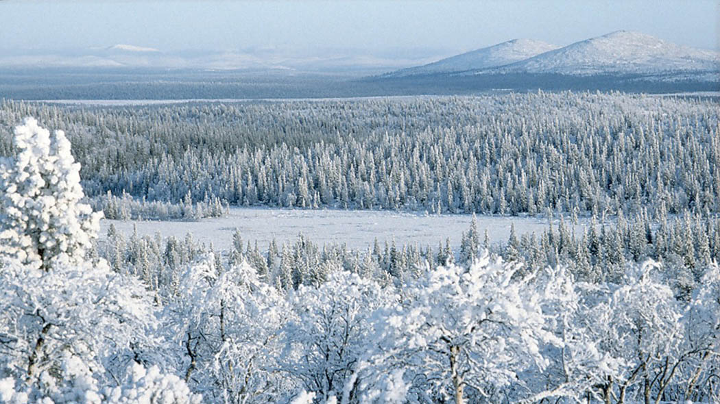 Lumiselta tunturin laelta alla avautuva maisema, jonka takana terävä huippuisia tuntureita.