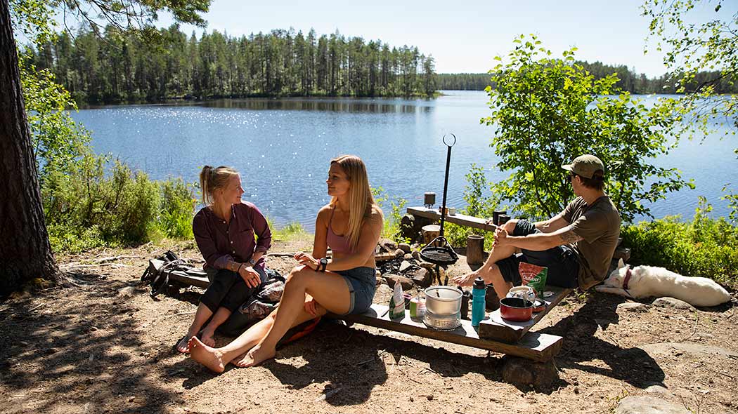 Ihmisiä istuskelemassa tulentekopaikalla. Taustalla kesäinen järvimaisema.